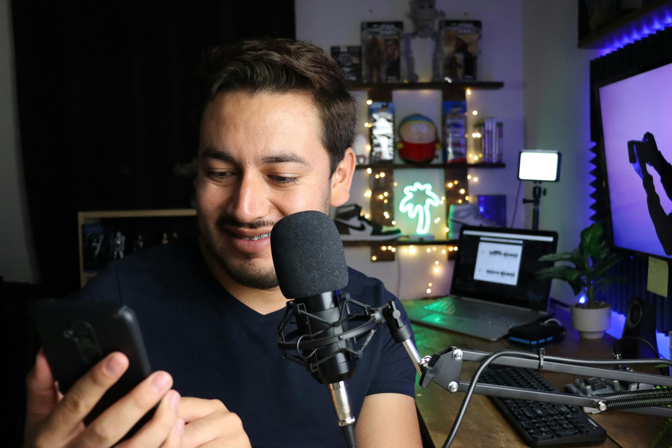 A man using a smartphone while seated at a desk with a microphone, surrounded by cozy decor.
